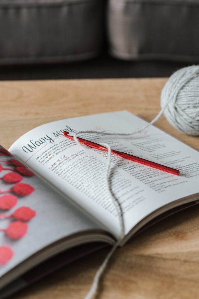 High angle of gray yarn ball with crochet hook and craft magazine on wooden table at home